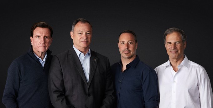 Four middle-aged men pose for the camera in front of a dark backdrop. From left to right, one wears a dark pullover,  one wears a dark suit with blue dress shirt, another wears a navy blue dress shirt, and the last one wears a white dress shirt.