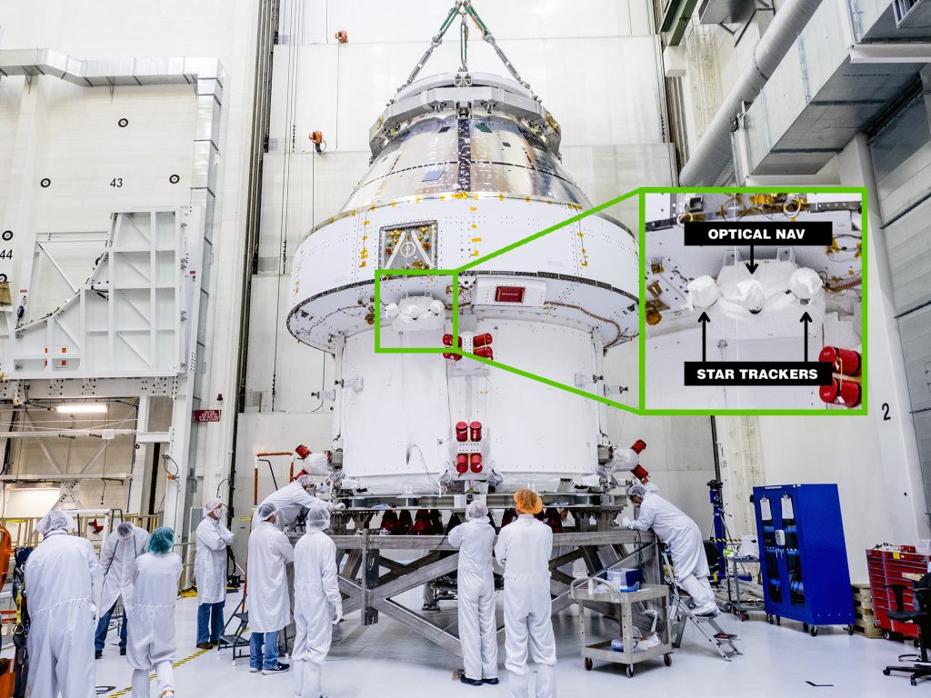 In a white clean room, employees dressed in white observe The Orion crew and service module stack for Artemis I was lifted out of the Final Assembly and Test (FAST) cell. The Orion star trackers are highlighted about halfway up the spacecraft.
