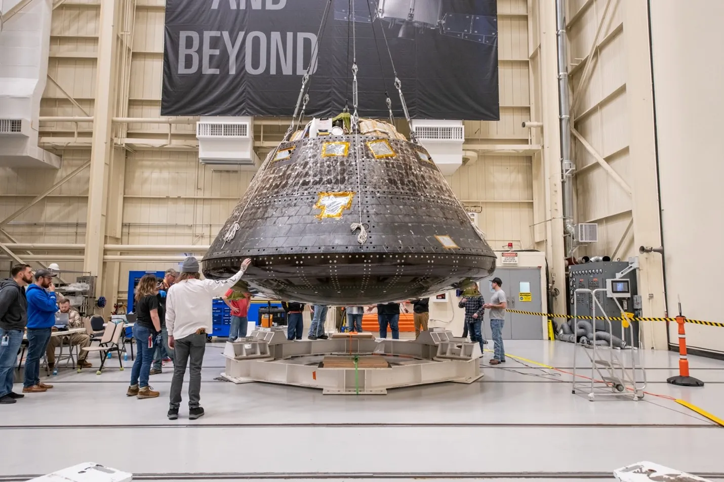 Inside the high-bay assembly area in the Space Environments Complex at NASA's Neil Armstrong Test Facility, the charred Orion capsule from Artemis I is hoisted about four feet above the round white metal framing that it will be tested in. Several engineers and technicians wearing jeans, casual shirts, and work boots surround the capsule.